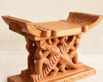 May include: A wooden stool with detailed carvings. The stool has a rectangular base, a carved central support, and a curved seat. The wood has a warm, natural tone, and the carvings depict figures and geometric patterns. The stool is made of wood.
