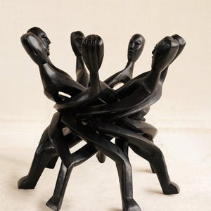 Puede incluir: Una escultura de madera tallada en color negro que representa a un grupo de figuras estilizadas de pie en círculo, con los brazos entrelazados. Las figuras tienen cuerpos lisos y alargados y rasgos faciales definidos, creando una forma continua.