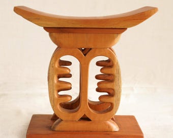 May include: A wooden stool with a light brown finish. The stool has a curved seat, a decorative central support with cut-out designs, and a rectangular base. The design is simple yet elegant, showing the natural wood grain. The stool is likely made from hardwood.