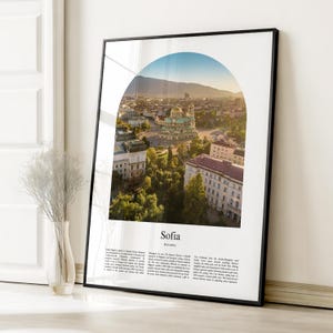 May include: A framed print featuring a cityscape of Sofia, Bulgaria, viewed through an arch. The image showcases the Alexander Nevsky Cathedral with golden domes, surrounded by buildings and greenery. The print is in a black frame.