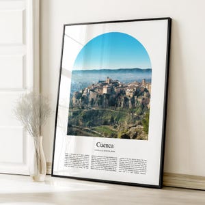 May include: Framed print of Cuenca, Spain, featuring a scenic view of the city nestled on a cliffside. The image is framed by a black border and includes a blue arch at the top. The print has the city's name and location at the bottom.