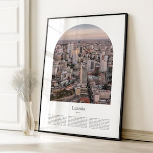 May include: A framed print featuring an aerial view of Luanda, Angola, seen through an arched window. The cityscape includes numerous buildings and skyscrapers. The print has a black frame and includes the city's name and descriptive text.