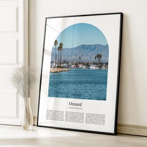 May include: Framed print featuring a scenic view of Oxnard, California, with a blue arch design. The image showcases a harbor with boats, palm trees, and mountains under a clear sky. The text "Oxnard, California, United States" is displayed.