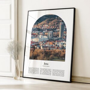 May include: A framed print featuring a cityscape of Jena, Germany, viewed through an arched window. The image showcases a mix of buildings, trees, and a tall skyscraper. The print is in a black frame and includes the city's name.