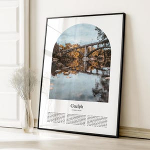 May include: A framed art print showing a bridge over a river, with the word "Guelph" and text below. The image is framed in a black border. The print is displayed against a white wall.
