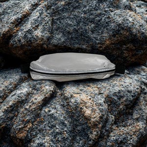 May include: A small, rectangular, light gray pouch with a black zipper and black fabric loops on each end. The pouch is resting on a rough, gray rock surface. The pouch appears to be made of a lightweight, possibly waterproof material.