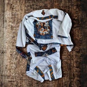 Op de afbeelding: Een witte kledingset met ingewikkelde blauwe en oranje patronen. De top heeft een centraal vierkant ontwerp met een vogelmotief. De set ligt op een houten oppervlak en toont het traditionele ontwerp.