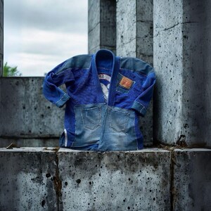 May include: A blue martial arts uniform jacket with denim accents. The jacket features a patchwork design with denim pockets and a leather patch. The background is a concrete structure under a cloudy sky.