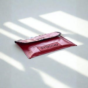 May include: A red leather clutch with the word "STÜCCIO" printed on a rectangular patch. The clutch has a black trim and is open, revealing a glimpse of the contents. The background is white with shadows.