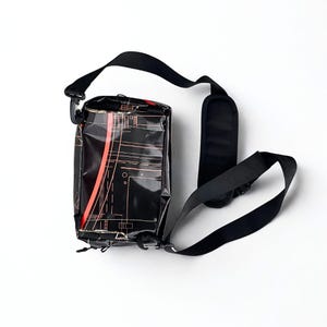 May include: Black crossbody bag with a geometric design in red and tan. The bag has a black adjustable strap and a padded shoulder rest. The bag is rectangular and has a buckle closure.