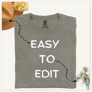May include: A sage green t-shirt with the words "EASY TO EDIT" in white block letters. The shirt is folded and has a tag on the collar. A brown hat and flowers are in the background.