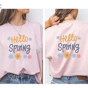 May include: Light pink t-shirt with the words "Hello Spring" in yellow and blue lettering, with flower graphics. The shirt is paired with blue denim jeans. The back of the shirt also displays the same design.