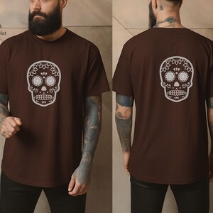 May include: Dark chocolate brown t-shirt featuring a white sugar skull design on the front and back. The skull has floral and dot details. The shirt is a short-sleeved crew neck.