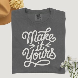 May include: A gray t-shirt with the words "Make it Yours" in white script. The shirt is folded and has a Comfort Colors tag. The image includes a tan hat and white flowers.