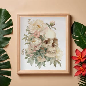 May include: A framed print featuring a human skull adorned with watercolor-style flowers. The skull is cream-colored, and the flowers are a mix of pink, cream, and green. The frame is a light wood color, and the overall aesthetic is a blend of gothic and floral.