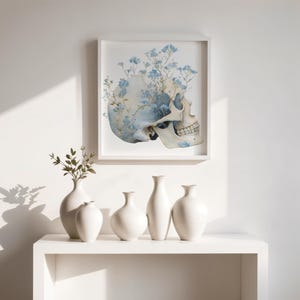 May include: A framed print featuring a watercolor illustration of a skull with blue flowers growing from it. Below, five white ceramic vases of varying shapes sit on a white shelf. The scene is set against a white wall.