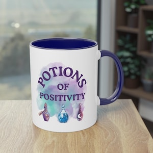 May include: White ceramic mug with a blue interior and handle. The mug features the text "POTIONS OF POSITIVITY" in purple, with three potion bottle illustrations in front of a watercolour background.