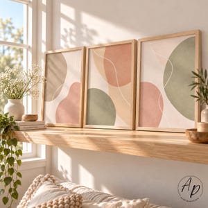 Peut inclure: Un ensemble de trois cadres muraux rectangulaires en bois clair, présentant des formes organiques abstraites dans des tons sourds de vert sauge, terre cuite et beige, rehaussées de fines lignes blanches fluides sur fond crème.