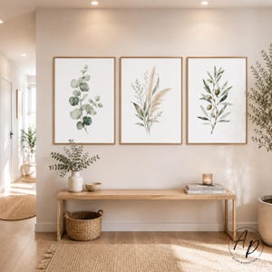 Op de afbeelding: Drie ingelijste botanische kunstprints met aquarel illustraties van eucalyptus, pampasgras en olijftakken. De prints hebben een neutraal kleurenpalet met tinten groen, beige en bruin, weergegeven boven een houten bank.