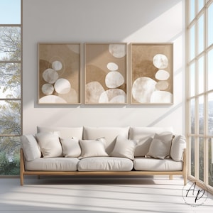 Könnte beinhalten: Drei gerahmte abstrakte Kunstdrucke mit beige-weißen organischen Formen. Das Kunstwerk hängt über einem cremefarbenen Sofa mit mehreren Kissen. Der Raum hat große Fenster und einen hellen Holzrahmen.