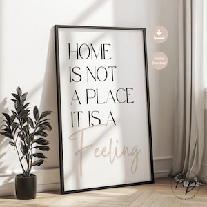 May include: A framed art print with the quote "HOME IS NOT A PLACE IT IS A Feeling" in black and beige lettering. The print is in a black frame and leans against a white wall. A potted plant is to the left of the print.