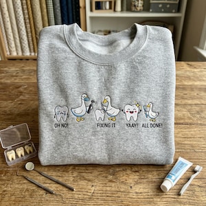 May include: A grey crew neck sweatshirt with a dental theme. The design features cartoon ducks and teeth with the phrases "OH NO!", "FIXING IT", "YAAAY!", and "ALL DONE!". Dental tools, toothpaste, and a toothbrush are also visible.