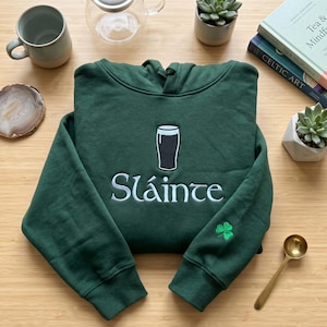 May include: A dark green hoodie with the word "Sl&aacute;inte" embroidered in white, along with a pint of stout and a green shamrock. The hoodie is folded on a wooden surface, surrounded by other objects.