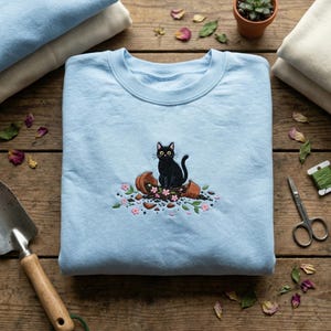 May include: A pale blue sweatshirt with an embroidered design of a black cat perched on a broken terracotta pot with pink flowers. The cat has yellow eyes and a curved tail. The sweatshirt is folded on a wooden surface.