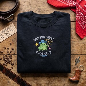 May include: Black sweatshirt with an embroidered design of a green frog in a cowboy hat holding a lasso. The text reads "Hold Your Horses Frog Club". A rodeo ticket and red bandana are in the background.