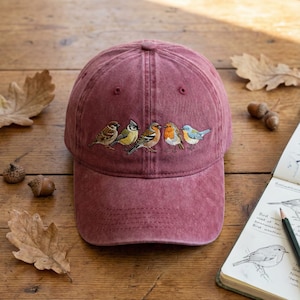 Peut inclure: Casquette de baseball bordeaux avec un motif brodé de cinq oiseaux colorés. La casquette est faite d'un matériau doux et texturé et possède une visière incurvée. Les oiseaux sont détaillés avec diverses couleurs, dont le marron, le jaune, l'orange, le rouge et le bleu.