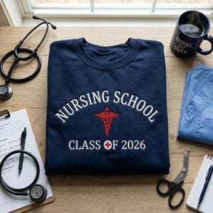 Peut inclure: Sweat-shirt bleu marine avec l'inscription blanche "NURSING SCHOOL", un symbole de caducée rouge et "CLASS OF 2026" en blanc. Un stéthoscope, une tasse et des fournitures médicales sont disposés autour du sweat-shirt.