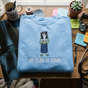 May include: A light blue sweatshirt with the embroidered phrase "WE SCAN AT DAWN". The sweatshirt features an embroidered frog holding an X-ray image. A mug with "RAD TECH" is visible.