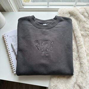 May include: Dark grey crew neck sweatshirt with an embossed design of an ultrasound machine, baby bottle, bow, and ultrasound probe. The sweatshirt is folded on a white surface next to a spiral notebook and a pen.