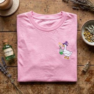 Könnte beinhalten: Hellrosa T-Shirt mit einer Cartoon-Gans, die einen lila Hexenhut trägt und eine grüne Zaubertrankflasche hält. Das T-Shirt ist auf einer Holzoberfläche gefaltet. Eine kleine Glasflasche mit dem Text "Witherbloom" ist sichtbar.