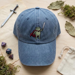 Könnte beinhalten: Blaue Denim-Baseballkappe mit einer gestickten Froschfigur, die einen roten Umhang trägt und ein Schwert hält. Die Kappe hat einen gebogenen Schirm und einen Stoffriemenverschluss. Das Bild zeigt auch Würfel und ein Spielzeugschwert.