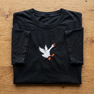 Peut inclure: T-shirt noir avec un motif d'oie blanche. L'oie est brodée avec des plumes blanches, des pattes orange et un bec orange ouvert. Le t-shirt est plié sur une surface en bois.
