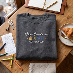 May include: A dark gray sweatshirt with the words "Chaos Coordinator COFFEE CLUB" in white script. The design includes a coffee cup, stars, a calendar, and a checkmark. A croissant and a notepad are also visible.