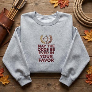 May include: A light gray sweatshirt with the embroidered phrase "MAY THE ODDS BE EVER IN YOUR FAVOR" in burgundy. The design includes a bow and arrow within a laurel wreath. The sweatshirt is displayed on a wooden surface with autumn leaves.