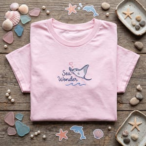 May include: A light pink t-shirt featuring an embroidered design of a cartoon stingray with the text "Sea Wonder" in blue script. The design includes a heart and blue waves. The shirt is displayed on a wooden surface with sea-themed decorations.