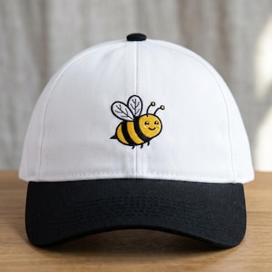 May include: A white baseball cap with a black brim, featuring an embroidered cartoon bee. The bee has yellow and black stripes, with a smiling face and antennae. The cap has a curved brim and a black button on top.