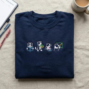 Peut inclure: Sweat-shirt bleu marine avec un motif brodé d'un chien dans diverses poses d'étude. Le chien est représenté en train de lire, de porter des livres, d'écrire et d'analyser un graphique. Le motif est blanc, noir et vert.