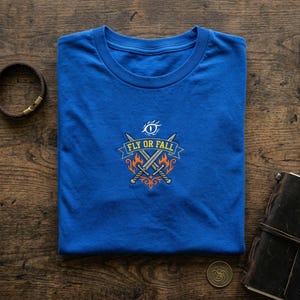 May include: A blue crew neck t-shirt with an embroidered design. The design includes the text "FLY OR FALL", crossed swords, flames, and an eye symbol. A leather bracelet and a notebook are also visible.
