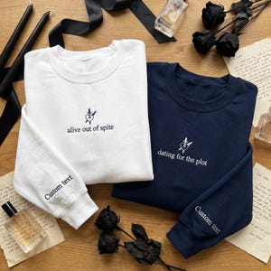 May include: Two crewneck sweatshirts, one white and one navy blue, with embroidered designs. The white sweatshirt says "alive out of spite" and the navy one says "dating for the plot." Both sleeves say "Custom text."