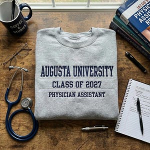 May include: A gray Augusta University sweatshirt with navy blue text reading "AUGUSTA UNIVERSITY CLASS OF 2027 PHYSICIAN ASSISTANT." The image includes a stethoscope, mug, glasses, books, and a notepad on a wooden surface.