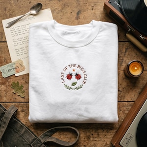 May include: A white crewneck sweatshirt with an embroidered design featuring two red ladybugs and the text "LAST OF THE BUGS CLUB" in a circular emblem. The design includes a star and green leaves. The sweatshirt is folded on a wooden surface.