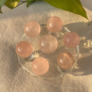 May include: Seven rose quartz spheres, each approximately 2.5 cm in diameter, are displayed on a clear glass plate. The spheres have a soft pink colour and a polished surface. The plate is set on a neutral-toned, textured surface.
