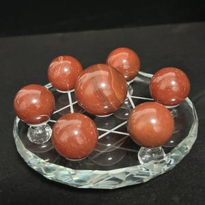May include: A collection of seven polished, spherical red jasper stones arranged on a circular, clear glass display stand. The stand features a geometric star pattern etched into the surface, with one larger sphere centered among six smaller spheres.