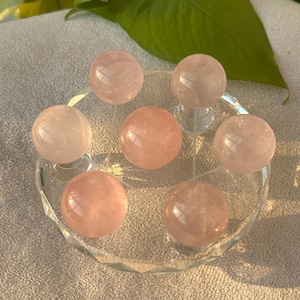 May include: Seven rose quartz spheres in shades of pink are displayed on a clear, star-shaped glass holder. The spheres have a smooth, polished surface. A green leaf is visible in the background.