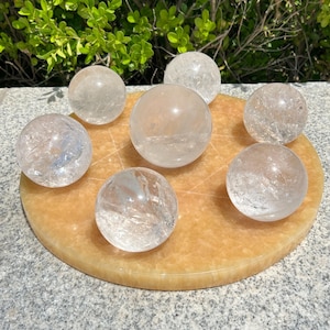 May include: Seven clear quartz crystal spheres displayed on a round, light orange-yellow stone base. The spheres vary in size and clarity, with internal patterns visible. The base features a subtle, star-like design. Green foliage is visible in the background.