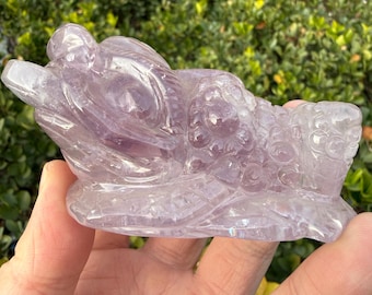 May include: A carved amethyst dragon figurine. The dragon is a light purple color and has detailed scales and features. The figurine is held in a hand against a backdrop of green foliage.